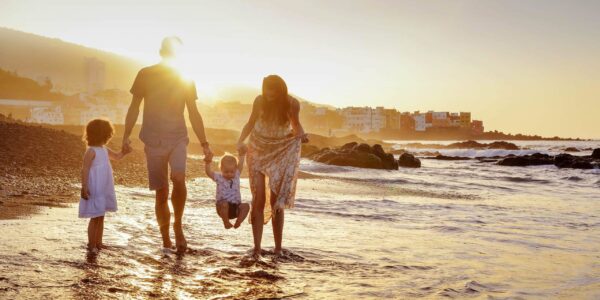 A family walking together on a beach as the sun sets in front of them, the man and woman are holding a young child between them as water laps around their ankles
